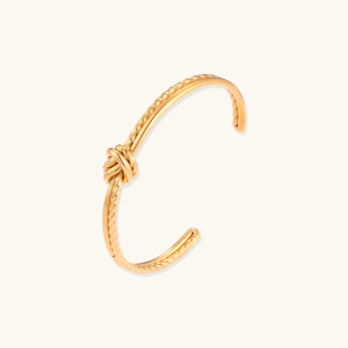 Gold knot cuff bracelet with a smooth and twisted band tied in a center knot, shown open on a cream background